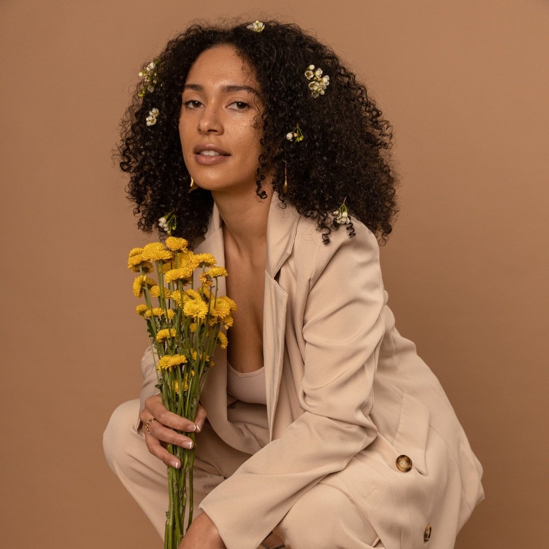 black female model in a loose fitting beige suit with curly hair and a bouquet of daisies