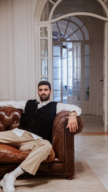 middle eastern man sitting on a brown leather couch in an old mansion with a gold watch 