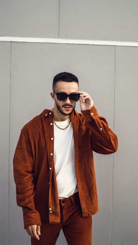 guy in a burnt orange coord set with shirt unbuttoned and white undershirt, wearing sunglasses and looking at the camera