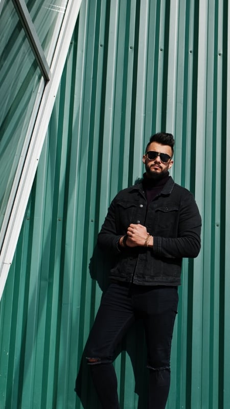 man in a full black outfit and sunglasses standing against a green wall