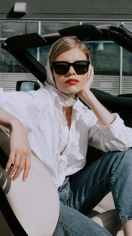 woman in a white shirt with a scarf around her head and wearing sunglasses, sitting in an open top car 
