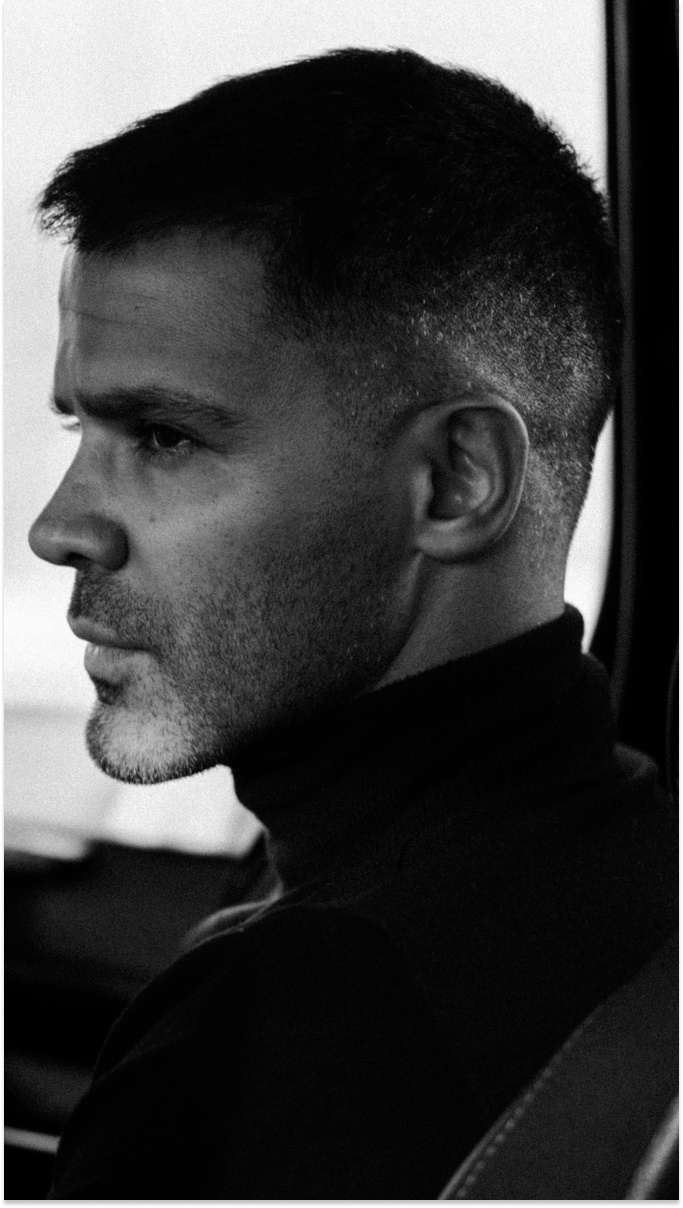 black and white side profile image of a fair man, with short hair and light stubble, wearing a polo neck