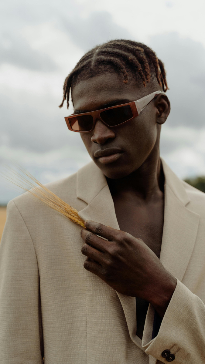 A brown skinned man with short cornrowed dreadlocks and a beige coloured suit, standing in a maize field, holding a strand of the agriculture against his shoulder