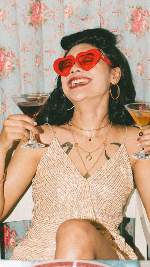 Model with black hair in a poof style, holding two martinis and wearing red, heart shaped sunglasses with a shimmery dress