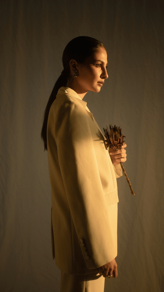A woman photographed from the side with her hair tied in a low ponytail, wearing a natural coloured suit, holding a single protea flower in her hand. She is standing in warm light cast from the sun setting or rising