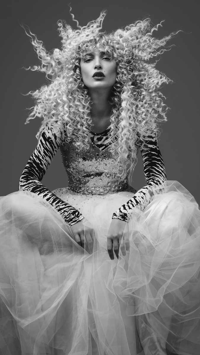 A woman with blonde, permed curly hair photographed in black and white in a long sleeve, zebra print top with a sequence vest layered over it and a tulle skirt. She is sitting down with her legs apart, hands draped on each leg and looking into the camera with a straight face