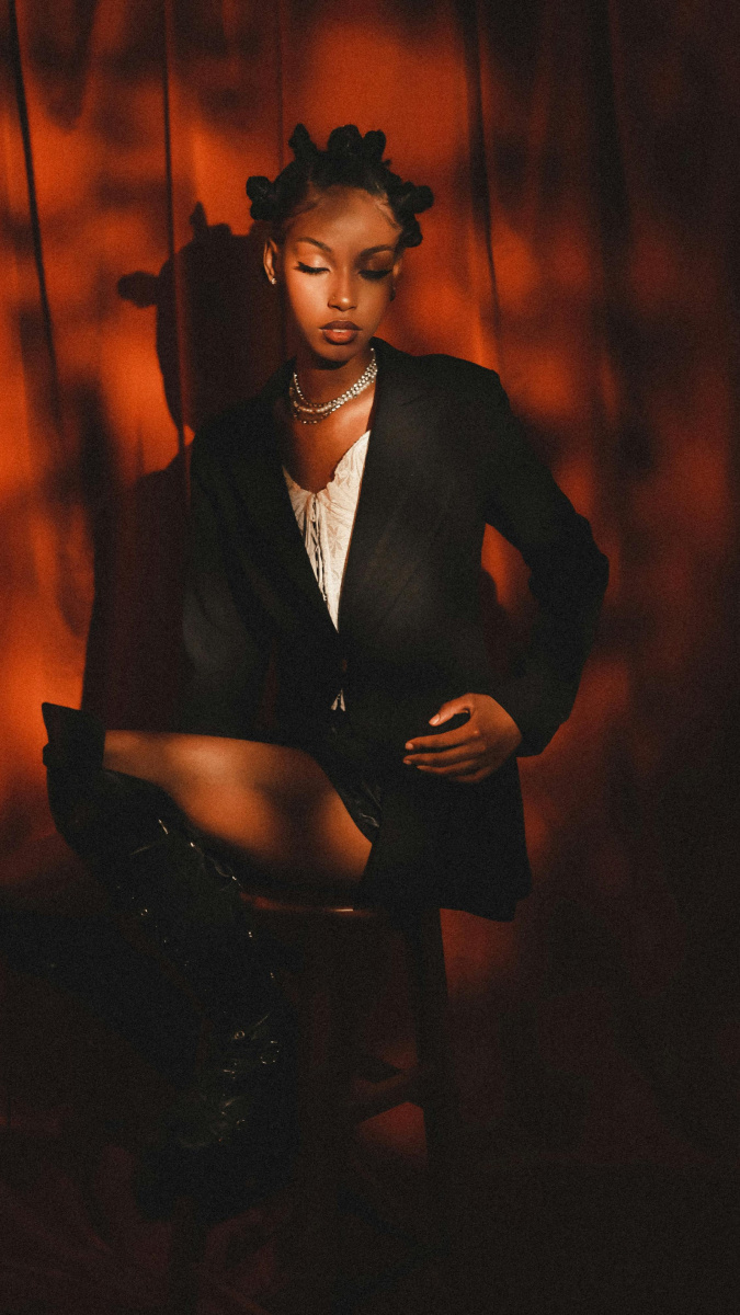 A woman with bantu knots in her hair, sitting on a small stool wearing a black blazer with a white corset underneath. She is looking downwards, away from the camera