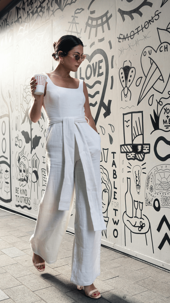 A woman with brown hair tied in a bun, wearing white linen pants and a white sleeveless top and round sunglasses, walking by a wall scattered with doodles, holding a coffee cup