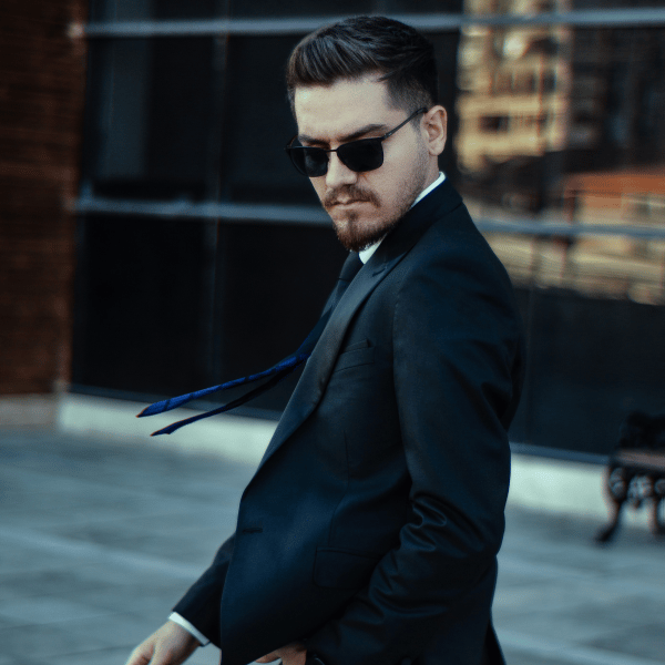 A light skinned man with a moustache and small beard in a black suit and blue tie, wearing black sunglasses photographed in motion, walking on a sidewalk with his face angled towards the camera and looking downwards