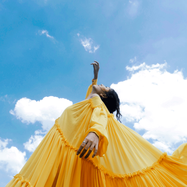 Image of a woman photographed from the ground angled upwards, wearing a long yellow dress with pleats and long sleeves, with one arm raised towards the sky