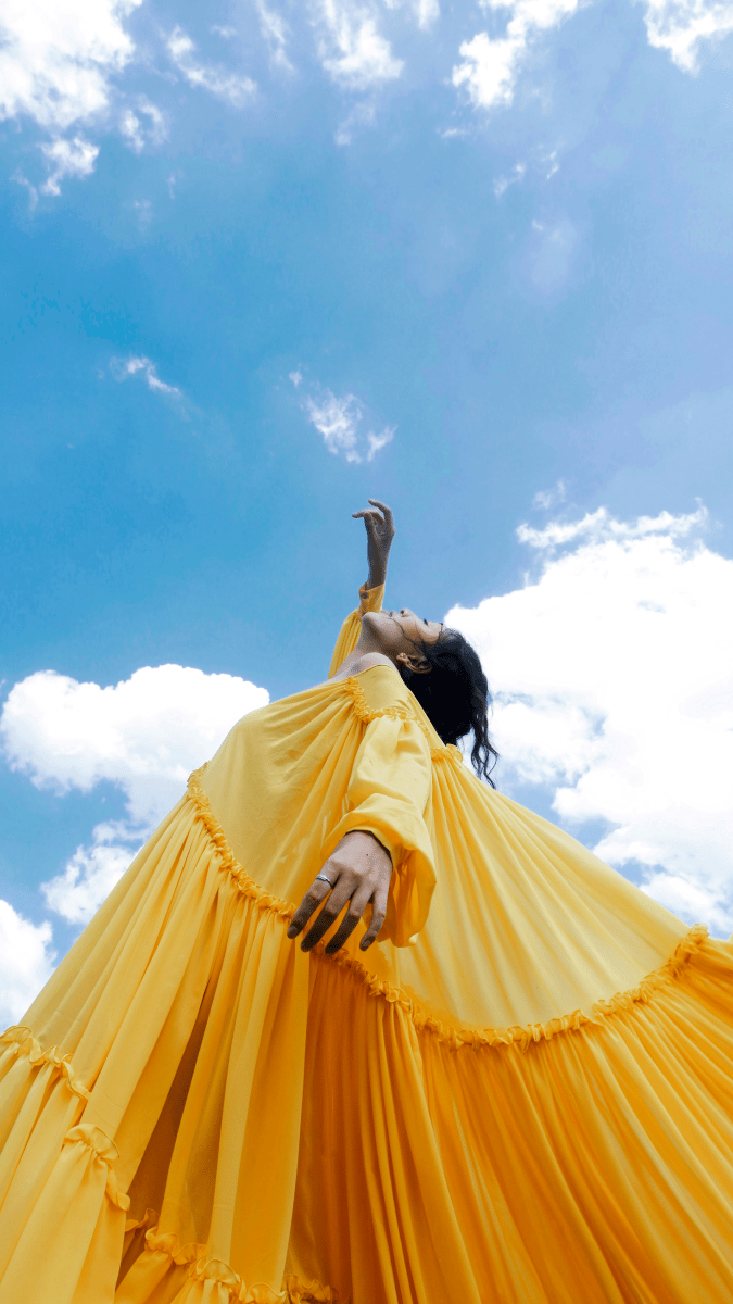 Image of a woman photographed from the ground angled upwards, wearing a long yellow dress with pleats and long sleeves, with one arm raised towards the sky