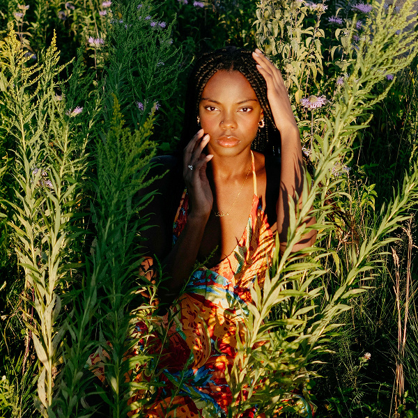 A woman in a floral dress, standing in tall grass and looking at the camera