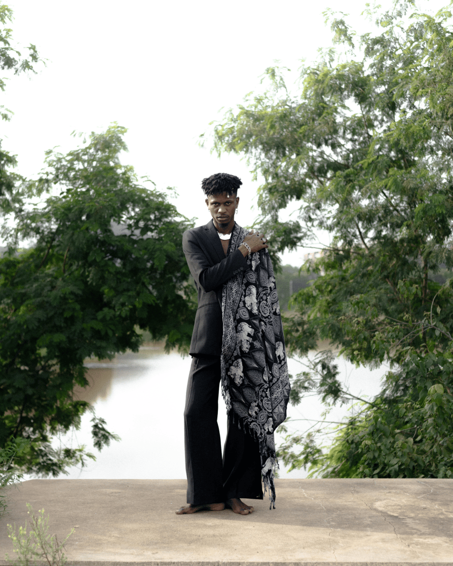 A young, barefoot man wearing flared black pants, a black blazer and chunky silver jewellery with a black and white tribal patterned piece of material over his left shoulder