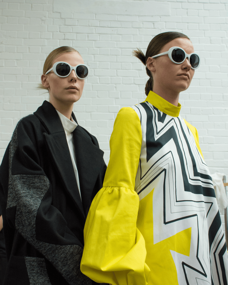 Two women, one with brunette hair and one with blonde, one wearing a yellow dress with geometric stripes and round white sunglasses, the other in a black coat and round, white sunglasses
