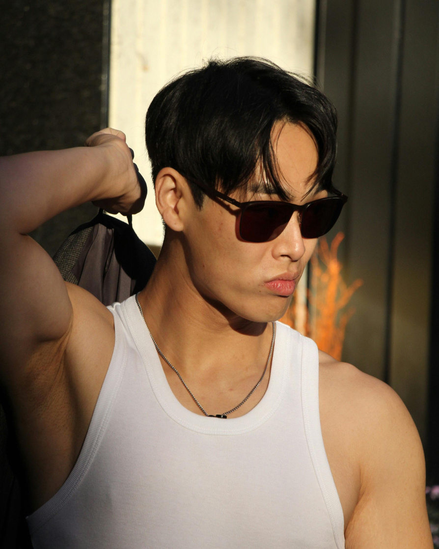 Asian model with short, dark straight hair, wearing square, brown sunglasses and a tight, white tank top, holding a blazer over his shoulder and looking down and to the right