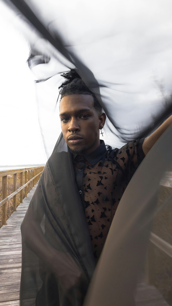 A dark skinned young man with his hair in a bun and looking into the camera, wearing a transparent, black, patterned shirt, draped in a satin shawl that blows in the wind