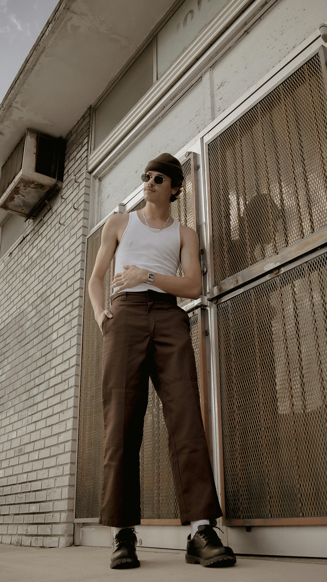 A young guy wearing a tight, white vest, with a brown beanie, sunglasses, brown formal pants and smart, black shoes