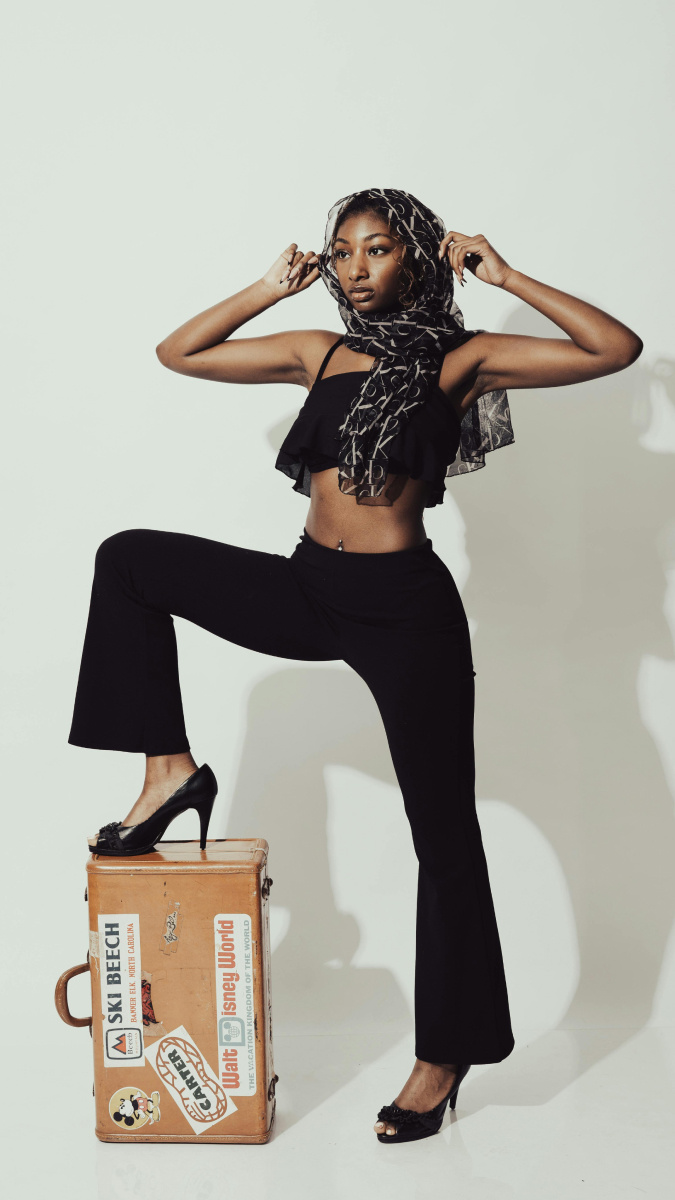 A dark skinned woman wearing a cropped black top, flared black pants, a scarf wrapped around her head and short, black heels, with one leg on top of an old, brown suitcase