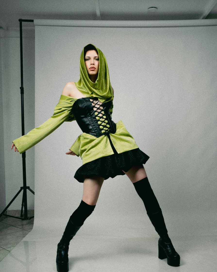 A model in a short lime green dress and a black skirt underneath, thigh high black boots, a corset over the dress and a loose green headwrap, photographed posing at the camera