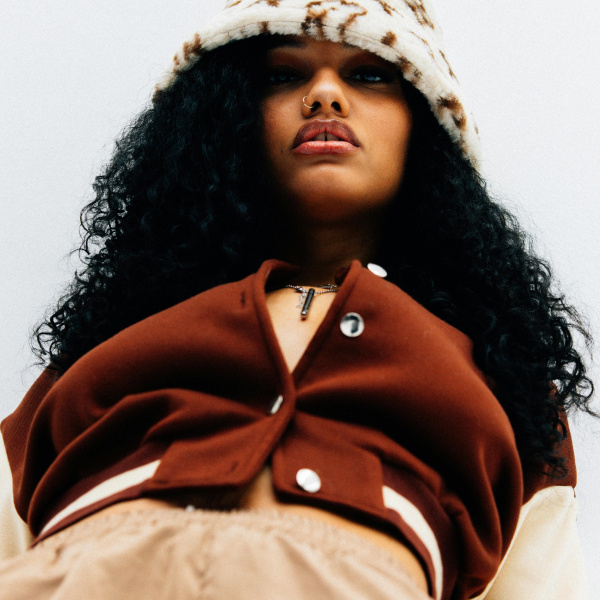 A photo of a brown skinned woman with black, curly hair, a nose piercing, a cow print wool hat and a maroon letterman jacket, looking down at the camera