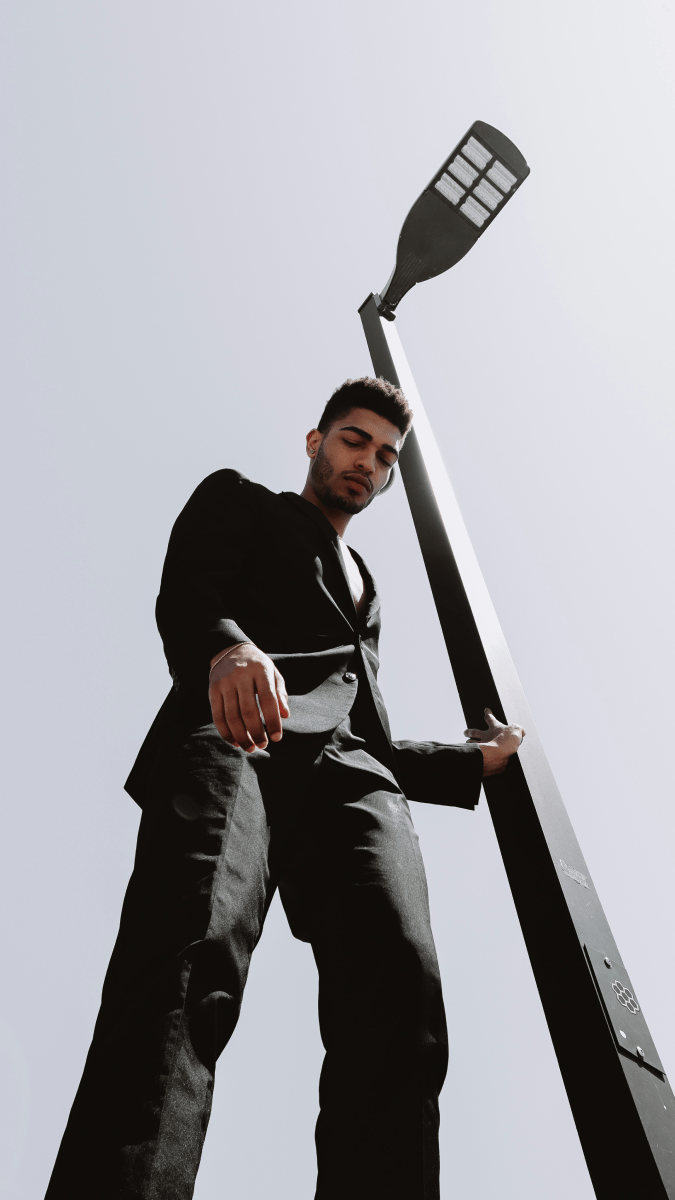 Young man in a black outfit with a formal black jacket, leaning against a light pole and looking down into the camera