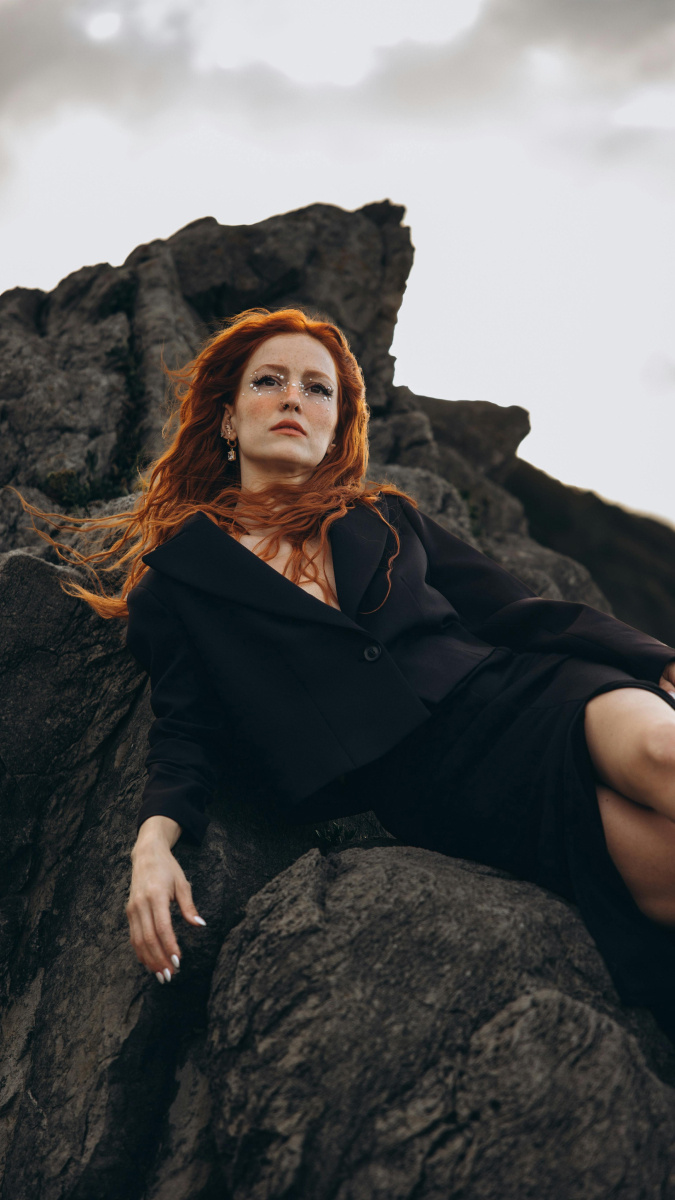 Red haired woman in a cropped, black blazer and tight black skirt, laying on a rock, staring into the distance