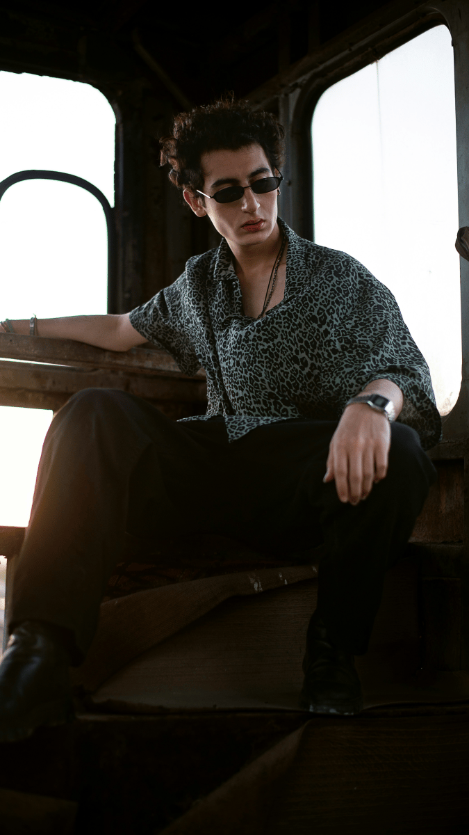 A young, light skinned man with medium length brown hair and black sunglasses sitting in a big piece of construction machinery wearing a short sleeve turquoise leopard print shirt and black pants. He is photographed looking to the right