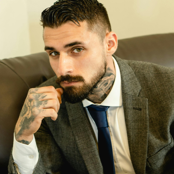 A young man with slick back brown hair and fair skin, wearing a textured suit and silk, dark blue tie, with tattoos on his neck and hand which he leans his face on