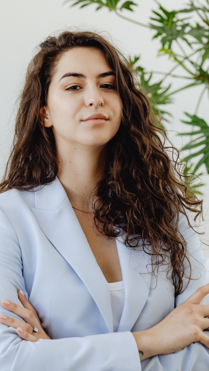 Curly, brown haired, light-skinned woman in a pastel blue suit with her arms crossed over her chest, smirking at the camera