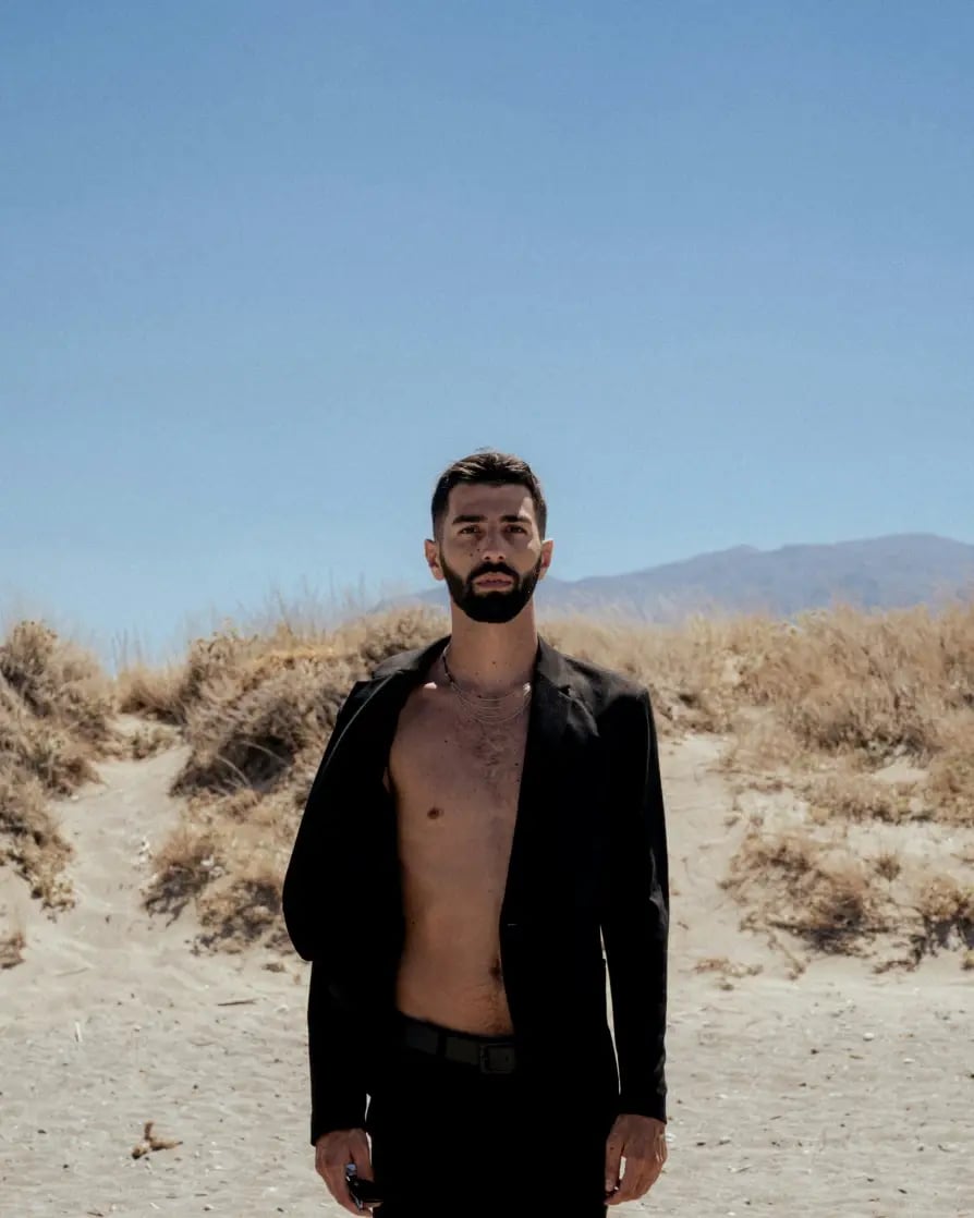 A young man with short, dark hair and a dark beard wearing an unbuttoned black linen shirt and standing in a dry, desert type area