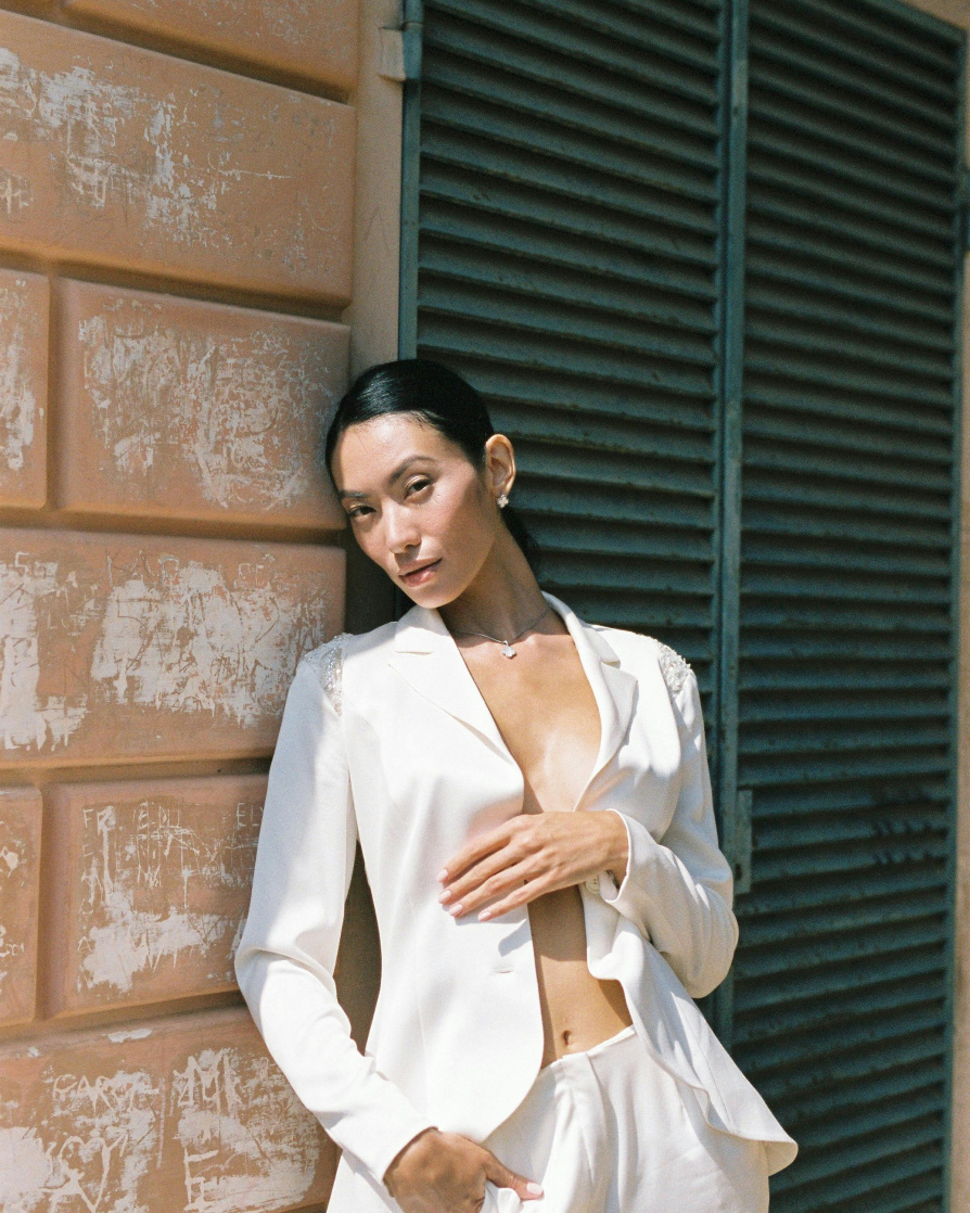 Asian model leaning against the wall wearing just a flowy white suit jacket and pants, with her hair slicked back into ponytail, looking at the camera