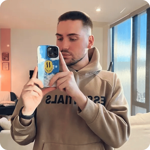 A young man with short hair, wearing a light brown hoodie and holding up a blue cloud covered phone case with a yellow smiley face