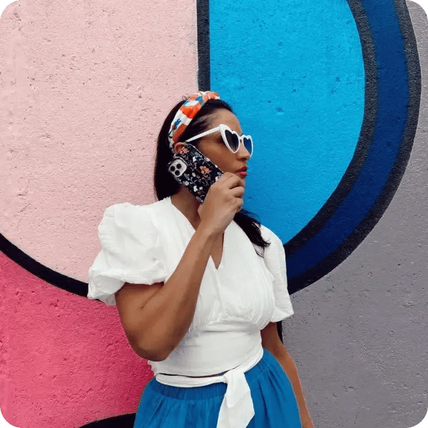A woman standing against a pink and blue graffitied wall, wearing white, heart shaped sunglasses, a white shirt and a blue skirt holding a black flower phone case to her ear