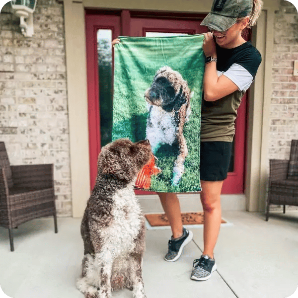 A woman holding up a blanket with her dog printed on it, and her dog sitting besides her looking up at her