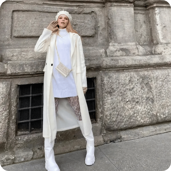 A woman with long blonde hair in a cream, knitted trenchcoat, wearing a thigh length polo neck with a cream beret, high white boots and an off white bag across her body