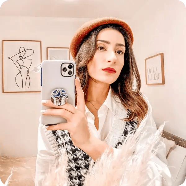 A woman with an orange beret, white shirt and knitted vest holding her phone with a custom designed popsocket