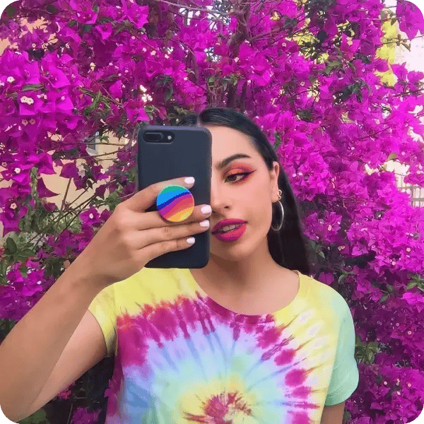 A girl with orange eyeshadow and red lipstick wearing a tie-dye shirt holding her phone up with a rainbow coloured popsocket