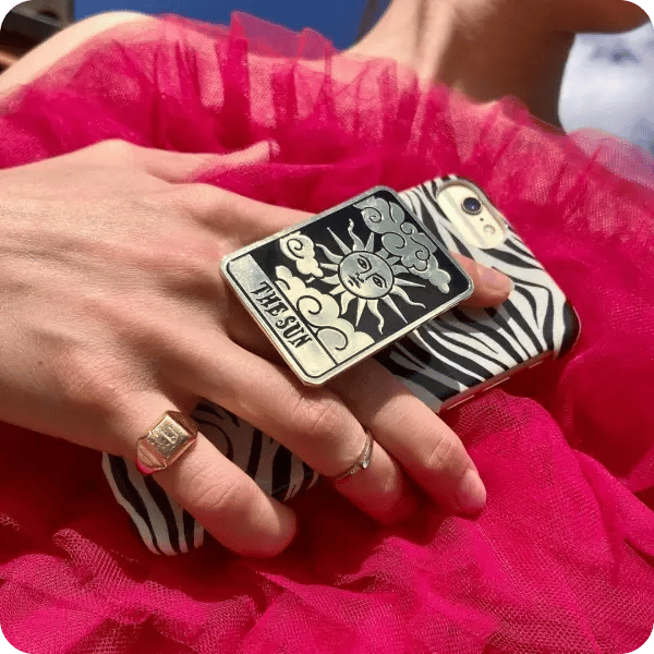 A close up of a woman in a pink tuiled dress, holding her phone with a zebra patterned cover and a sun tarot styled popsocket