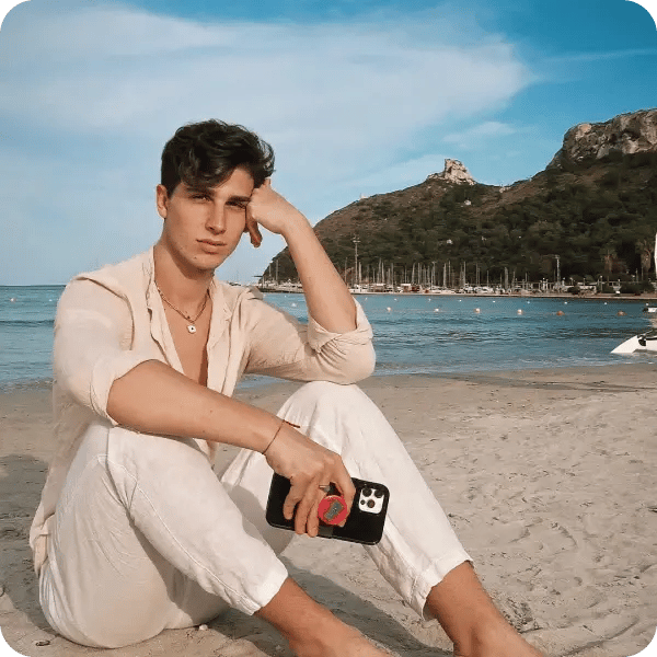 A young man with light brown hair, sitting on the beach in a linen shirt and pants, holding his phone with a red popsocket
