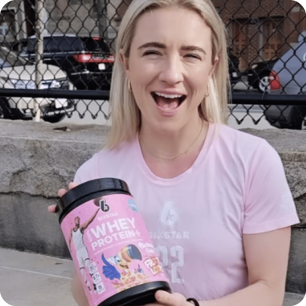 A women in a pink shirt holding a tub of whey protein in her hand and smiling at the camera