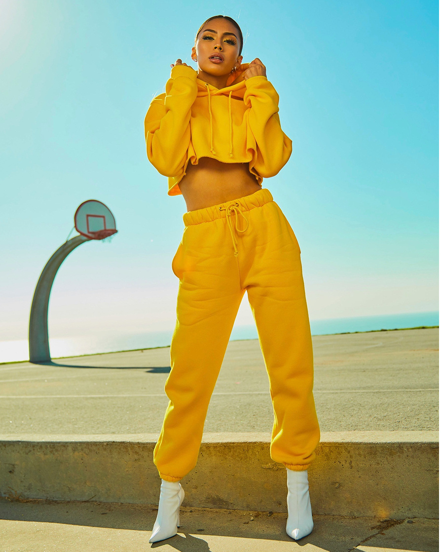 Female model wearing a cropped yellow hoodie and yellow sweatpants with a slicked back bun, standing at the edge of a basketball court and looking into the camera