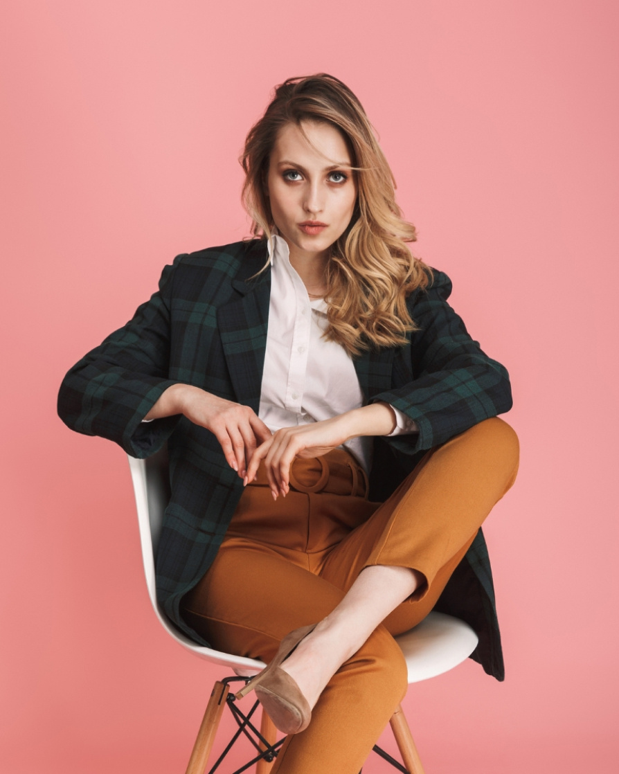 A woman in a green and navy blue checkered blazer with brown pants, pointy brown heels and dark blonde hair sitting in a chair, looking at the camera