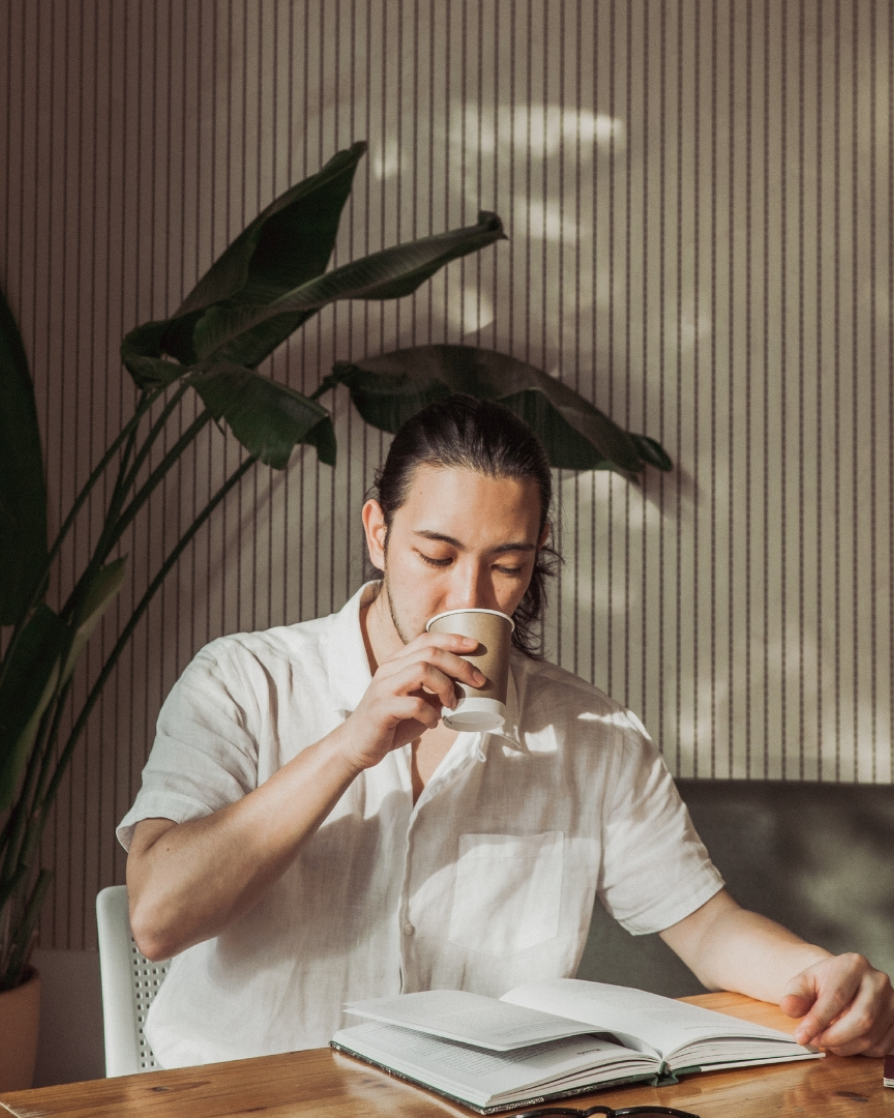 image of a young, asian man in a cafe with wavy brown hair tied into a pony tail, wearing a linen shirt, sipping on a coffee