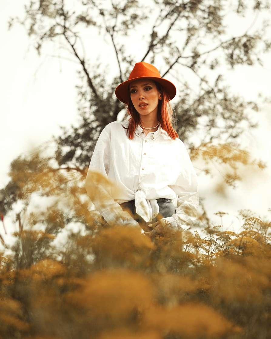 A woman standing in the middle of a field, with her upper half visible, wearing a white shirt and a orange brown hat with red hair, looking away from the camera