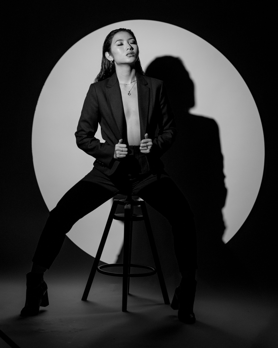 An asian woman in a suit, with her hands holding each side of the lower part of the suit opening, wearing nothing underneath the jacket with her hair slicked back, eyes closed and photographed in black and white with her head tiled backwards